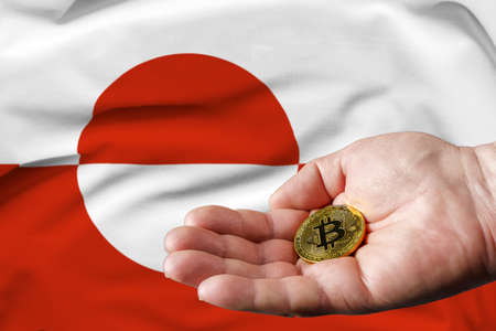 Golden bitcoin coin in man's hand, Greenland flag in the background.の写真素材