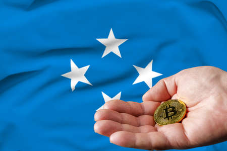 Golden bitcoin coin in man's hand, Micronesia flag in the background.