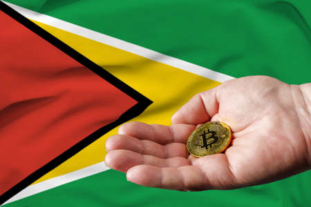 Golden bitcoin coin in man's hand, Guyana flag in the background.の写真素材