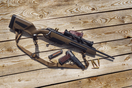 Scout bolt rifle, APS pistol, knife and cartridges on a wooden platform. View from above