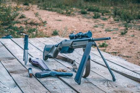 Scout bolt rifle, APS pistol, knife and cartridges on a wooden platform. Side view