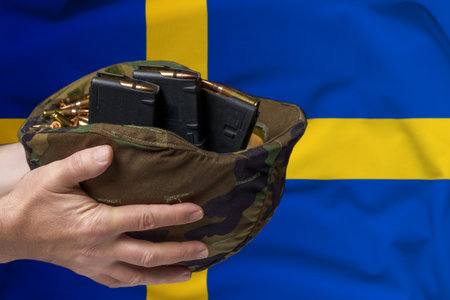 A military helmet with cartridges and magazines for a rifle in the hands of a man against the background of the flag of Sweden. The concept of selling weapons or military assistanceの写真素材