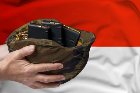 A military helmet with cartridges and magazines for a rifle in the hands of a man against the background of the flag of Indonesia. The concept of selling weapons or military assistanceの写真素材