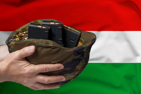 A military helmet with cartridges and magazines for a rifle in the hands of a man against the background of the flag of Hungary. The concept of selling weapons or military assistanceの写真素材