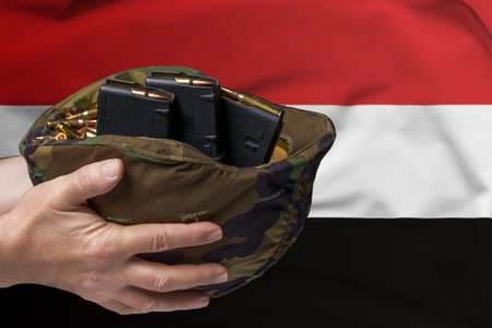 A military helmet with cartridges and magazines for a rifle in the hands of a man against the background of the flag of Yemen. The concept of selling weapons or military assistanceの写真素材