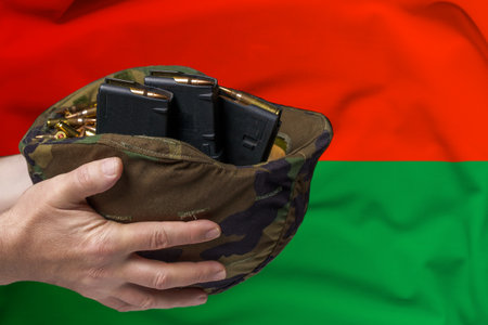 A military helmet with cartridges and magazines for a rifle in the hands of a man against the background of the flag of Burkina Faso. The concept of selling weapons or military assistanceの写真素材