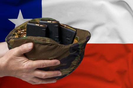 A military helmet with cartridges and magazines for a rifle in the hands of a man against the background of the flag of Chile. The concept of selling weapons or military assistanceの写真素材
