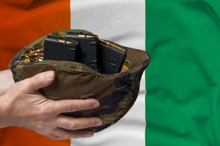 A military helmet with cartridges and magazines for a rifle in the hands of a man against the background of the flag of Ivory Coast. The concept of selling weapons or military assistanceの写真素材