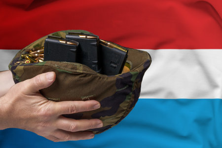A military helmet with cartridges and magazines for a rifle in the hands of a man against the background of the flag of Luxembourg. The concept of selling weapons or military assistanceの写真素材