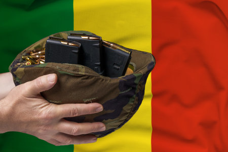 A military helmet with cartridges and magazines for a rifle in the hands of a man against the background of the flag of Mali. The concept of selling weapons or military assistanceの写真素材