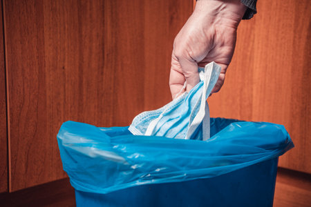 A man throws a medical mask into the trash. Man's hand with a medical mask and a trash can in blue.の写真素材