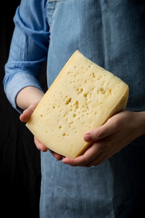 Woman holding slice of cheese wearing blue apron on dark background. Studio shot