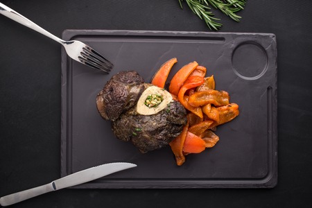 Cooked osso buco steak with grilled bell pepper on stone board on black background top view