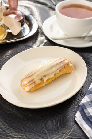 Homemade eclair dessert with vanilla sauce on white plate with cup of tea on the table side view