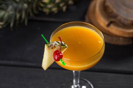yellow alcohol cocktail with pineapple and cherry in martini glass on black wooden table side viewの写真素材