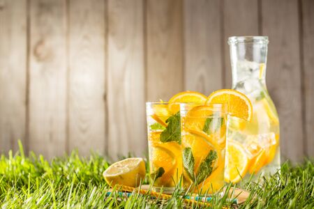 cold lemonade drink with orange and mint in tall glass on green grass lawn wooden background side viewの写真素材