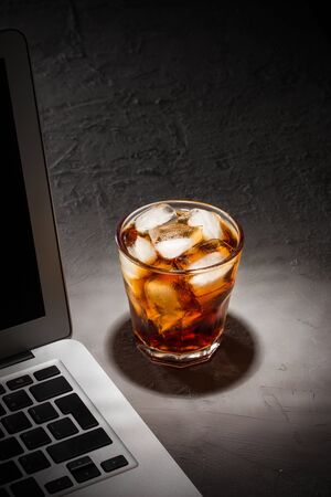 glass of cola with ice and laptop on grey stone table on black background side viewの写真素材