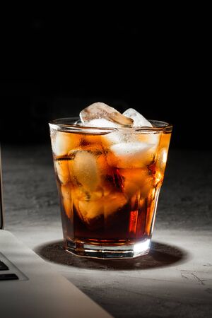 glass of cola with ice and laptop on grey stone table on black background side viewの写真素材