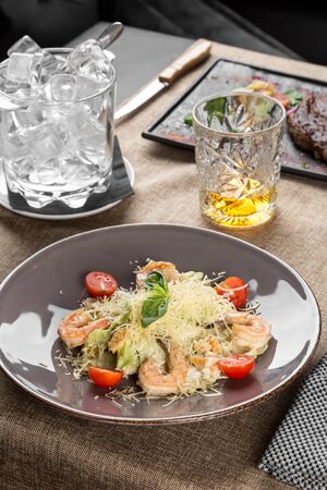 Healthy Grilled Caesar Salad with shrimp, parmesan Cheese and tomatoes and glass of whiskey on the table at restaurant side viewの写真素材