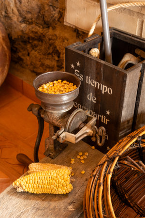 A rustic, antique hand-crank corn grinder filled with dried corn kernels, sitting on a wooden surface next to an ear of corn. In the background, a wooden crate and a woven basket suggest a traditional, harvest-time setting. This image evokes concepts of vintage farming, natural food preparation, and agricultural heritage.の写真素材