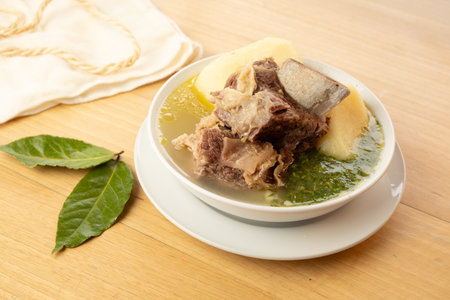 A hearty bowl of traditional Colombian Caldo de Costilla (beef rib soup) featuring tender chunks of beef rib and large pieces of potato, served in a clear savory broth. The soup is garnished with fresh herbs, likely cilantro or green onion. Next to the bowl, fresh bay leaves and a rustic cloth napkin complete the natural, home-cooked feel on a light wooden table, highlighting this popular comfort food.の写真素材