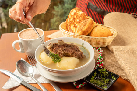 A hearty bowl of traditional Colombian Caldo de Costilla (beef rib soup) featuring tender chunks of beef rib and large pieces of potato, served in a clear savory broth. The soup is garnished with fresh herbs, likely cilantro or green onion. Next to the bowl, fresh bay leaves and a rustic cloth napkin complete the natural, home-cooked feel on a light wooden table, highlighting this popular comfort food.の写真素材