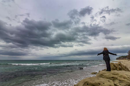 A woman on the top of a rock enjoys the view of sea and cloudy skyの写真素材