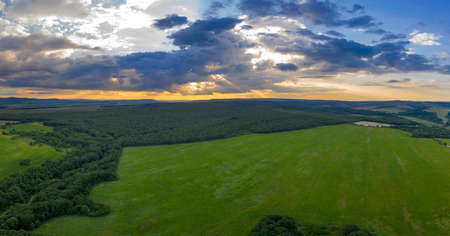 Amazing panoramic view of cloudy sky with sun rays over green field and forestの写真素材