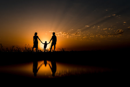 Silhouettes of Happy family holding hands with reflection on backdrop of the sea, sun, clouds in fiery red, orange colors.の写真素材