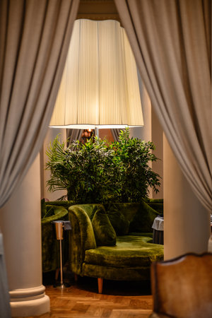 An elegant lounge area featuring a luxurious green velvet sofa and vibrant indoor plants, softly illuminated by a large floor lamp positioned between two columns. The space is framed by neutral curtainsの写真素材