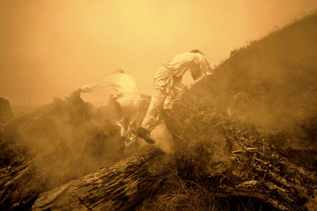 Two people in flame retardant suits climb into burning forest. Two firefighters make their way through smoke. warm tones.の写真素材