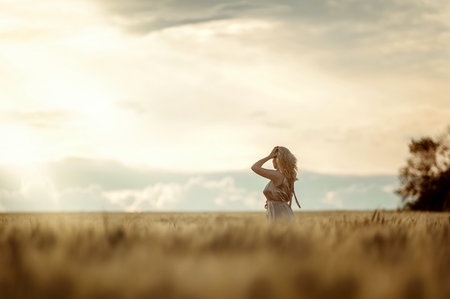 Profile portrait of a blonde girl in a golden dress, holding her hand to her head, standing in a wheat field against the background of the evening sky. Country style.の写真素材