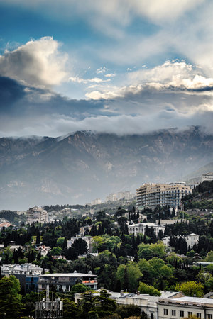 Cityscape with modern buildings, lush greenery, and mountains under dramatic cloudy sky during day, no people. Scenic view blending urban life with natural beauty.の写真素材