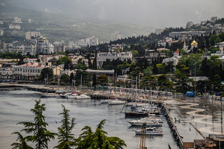 Coastal city with marina of yachts, modern buildings, and mountains under overcast sky after rain,...