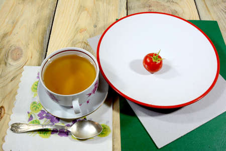 A cup of tea filled with tea is framed on a wooden table with a red tomato on a white plate.の写真素材