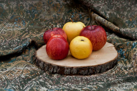 Three red and two yellow organic apples lie on a board cut from a thick tree branch. The background of the photo is a piece with a beautiful ornamentの写真素材