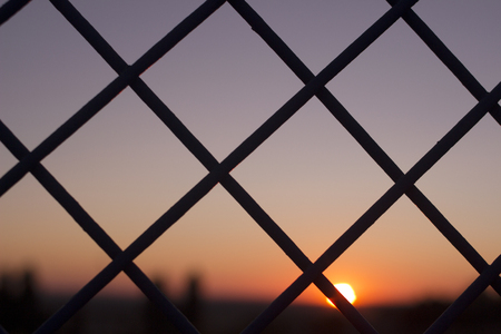 Horizontal color digital photo of setting evening sun and red and blue sky shining through a metal wire mesh fence in silhouette at dusk in dark tones with bokeh blur shallow depth of focus defocused landscape background.の写真素材
