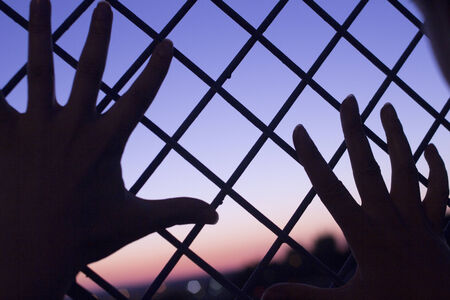 Color digital photo of setting evening sun and red and blue sky shining through a metal wire mesh fence with two human hands of a man against the fence with outstretched fingers in silhouette at dusk in dark tones with bokeh blur shallow depth of focus.の写真素材
