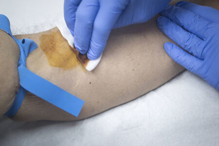 Close-up color photo of a female hospital clinic nurse wearing sterile blue gloves and senior male patient aged 65-70 giving a blood donation sample from the interior of his elbow resting arm on sterile blue white defocused background.の写真素材