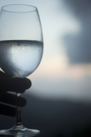Water glass held in hand by man in silhouette with sky and clouds behind defocusedの写真素材
