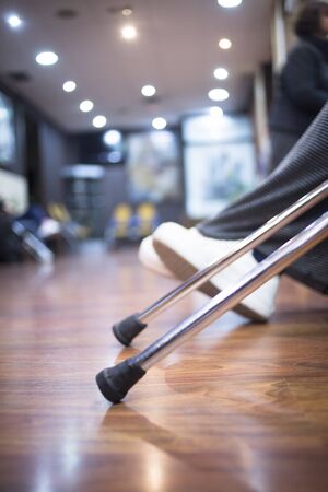 Aluminium metal crutches on floor in hospital clinic waiting room with legs defocused behind.