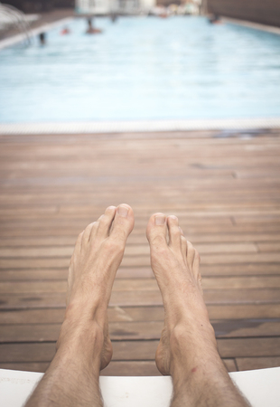 Legs and feet of man sunbathing on sun lounger in summer by swimming pool and wooden sundeck.