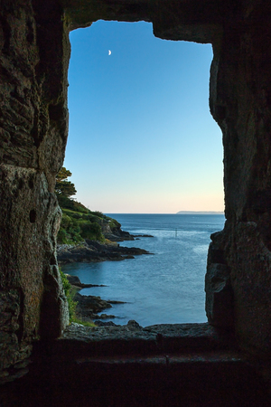 View of Polruan Harbour through the Castle, Cornwall, looking out onto blue sea and small moon.の写真素材