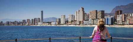 City scape panorama - lady gazes over blue sea to city backdrop and skyscrapers. Playa de Levante, Benidorm, Costa Blanca, Spain.の写真素材