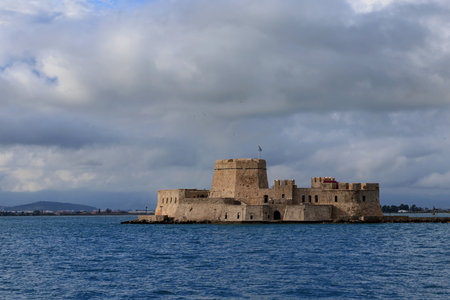 Bourtzi Castle in Nafplio, Greece, surrounded by sea and mountains under a moody sky with layered...