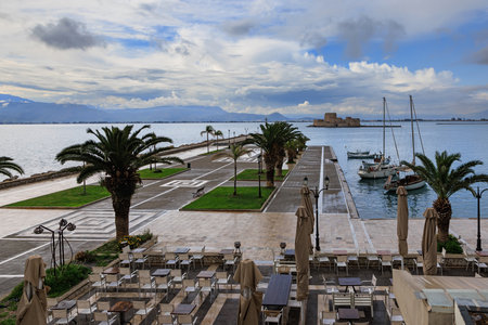 Scenic waterfront promenade in Nafplio, Greece with palm trees, empty cafÃ© terrace, yachts, and view of Bourtzi fortress under dramatic sky.の写真素材