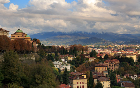 Elevated view of Bergamo, Italy, featuring terracotta rooftops, fall foliage, a church tower, and distant mountains under dramatic clouds.の写真素材
