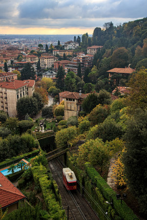 A red funicular climbs through lush greenery and fall foliage in Bergamo, Italy, surrounded by hillside villas and city rooftops under a cloudy sky.の写真素材