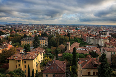 Elevated view of Bergamo, Italy, featuring terracotta rooftops, fall foliage, a church tower, and distant mountains under dramatic clouds.の写真素材