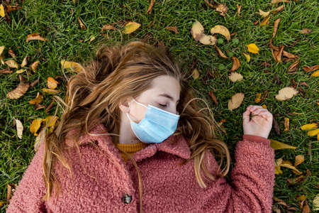 Concept of blond teen girl laying on her back in grass with a face mask on and her eyes closed. Tired of Covid virus.の写真素材
